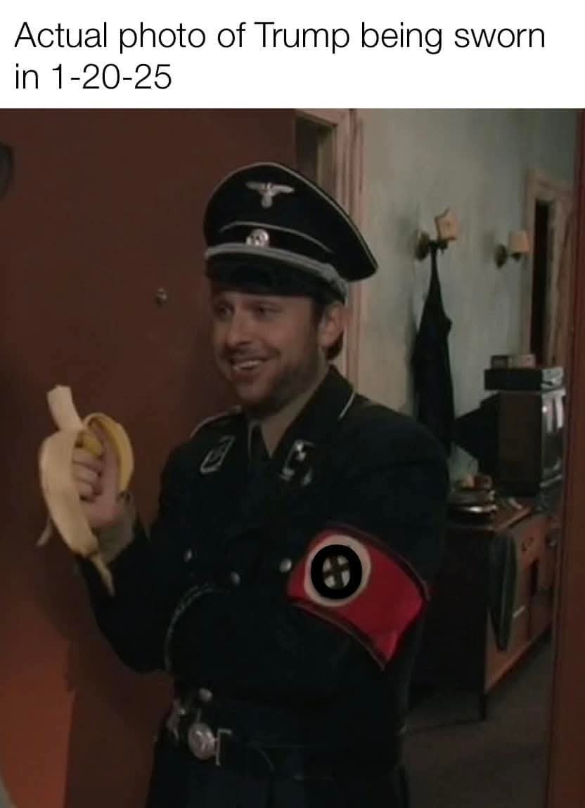 A man in a dark uniform with a Nazi-style cap and armband holds a peeled banana while smiling. The armband has a red…