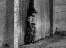 A black-and-white photo shows a young girl standing in a narrow, dilapidated doorway, looking out. Her face is obscured by a…