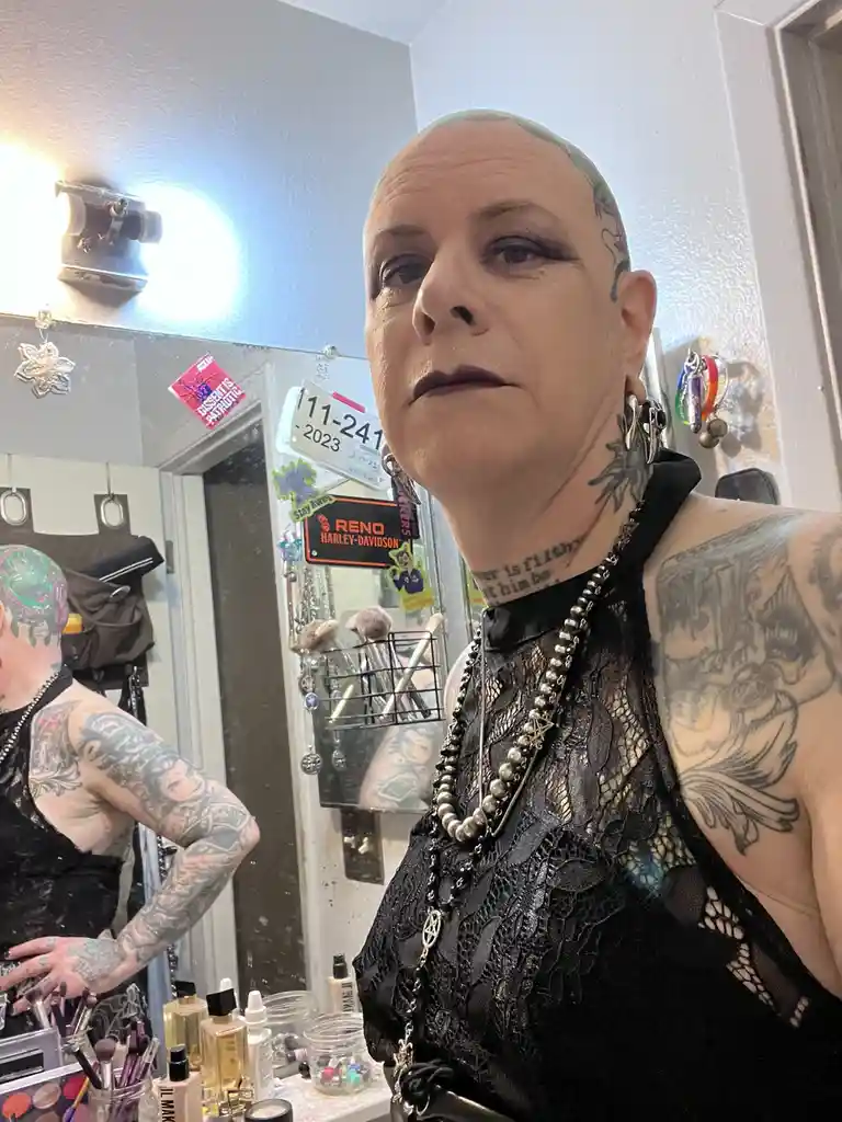 A person with a shaved head, heavy makeup, and numerous tattoos poses in front of a mirror in what appears to be a dressing…