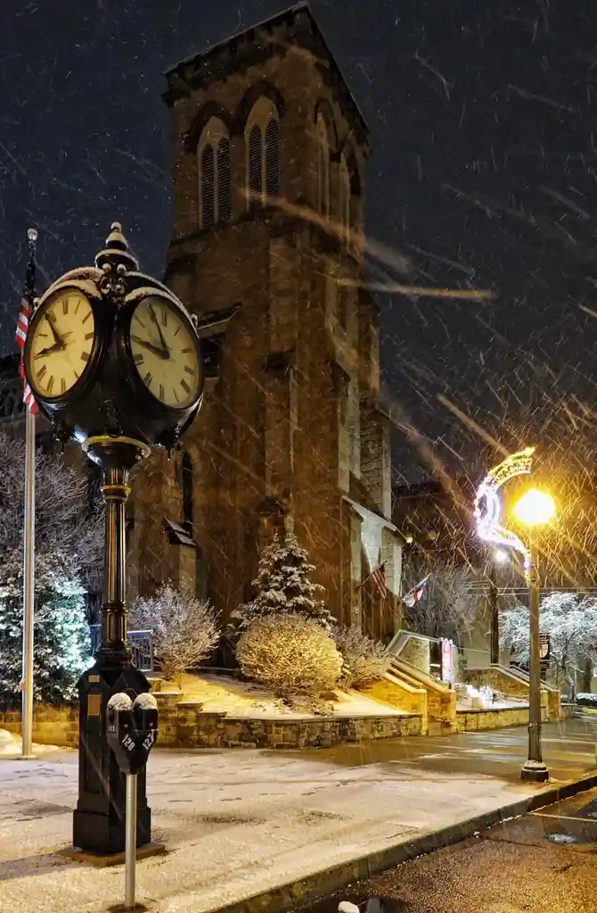 A snowy night scene features a vintage double-faced street clock in the foreground, its hands pointing to 11:59. Behind it…