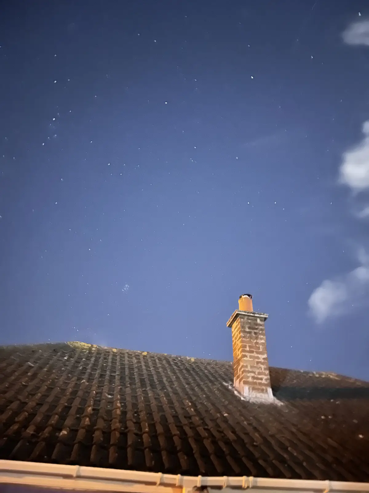 A low-angle nighttime photo shows a dark tiled roof with a brick chimney against a starry blue sky with faint clouds. The…