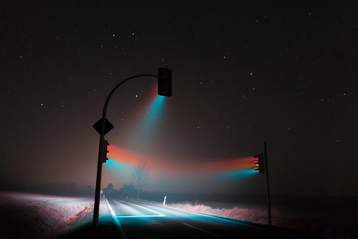A nighttime photograph shows two traffic lights on poles casting vibrant, colorful beams of red, orange, and blue light…