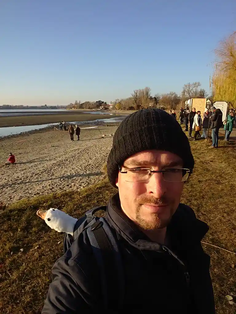 A man in a black beanie and glasses takes a selfie outdoors near a beach, with a small white duck peeking out from behind…