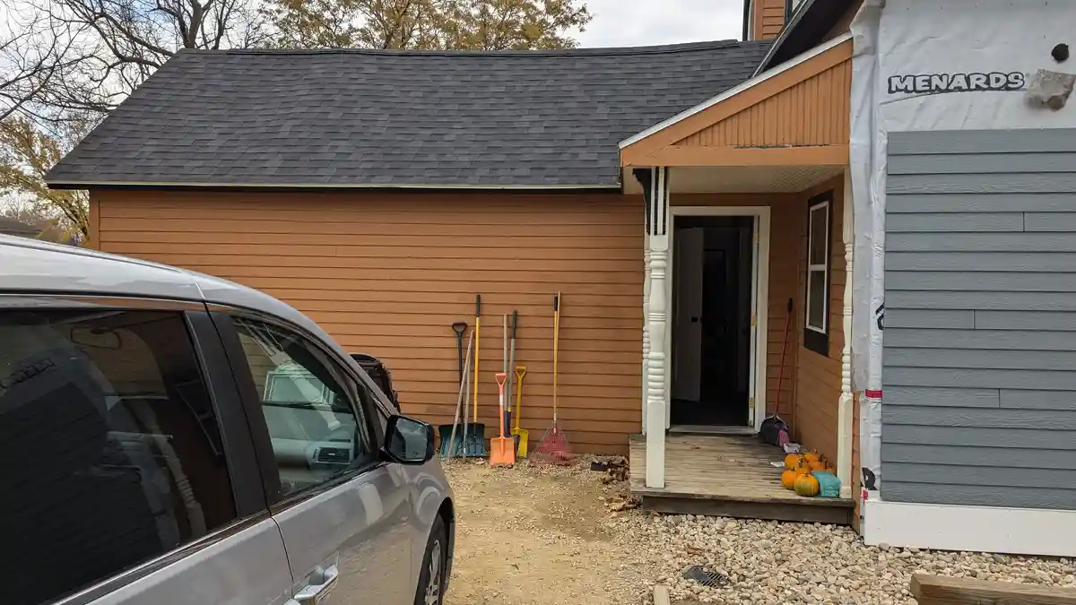 A house with orange siding and a gray roof has a section covered in gray siding and white Menards construction wrap. A…