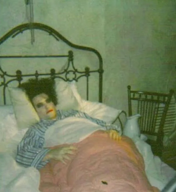 A person with dark curly hair and red lipstick lies in bed under a pink blanket, wearing a striped shirt, with a metal…
