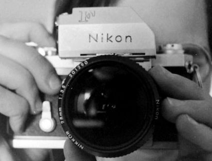 A black and white photo shows a person's hands holding a Nikon film camera with a 28mm lens. The camera's top plate has…