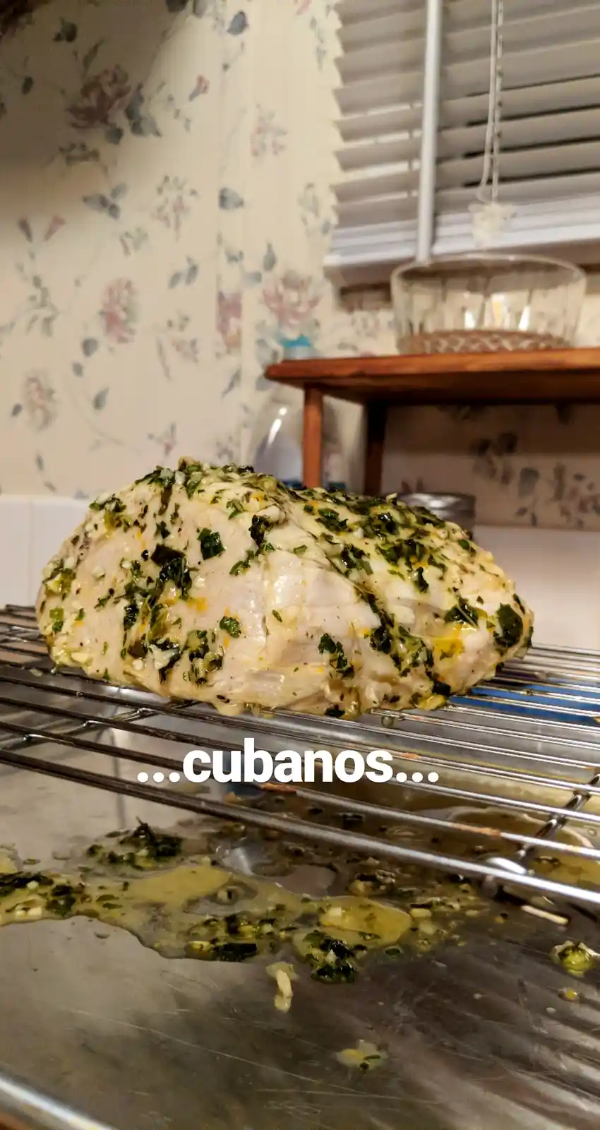 A large, herb-covered piece of cooked chicken breast sits on a metal cooling rack over a baking sheet with drippings. The…