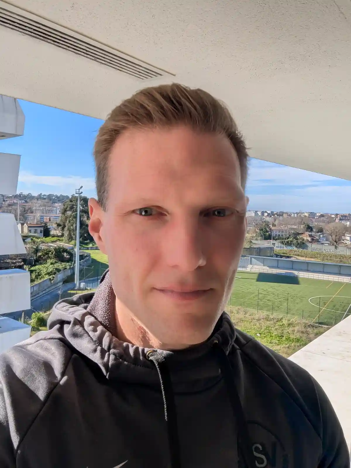 A man in a dark hoodie takes a selfie from a balcony overlooking a green soccer field and cityscape under a blue sky. He has…