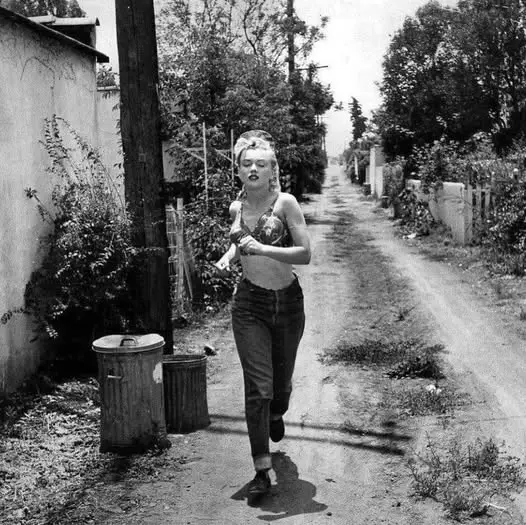 A black-and-white photo shows a woman with blonde hair running down a narrow, unpaved alleyway. She is wearing a halter top…