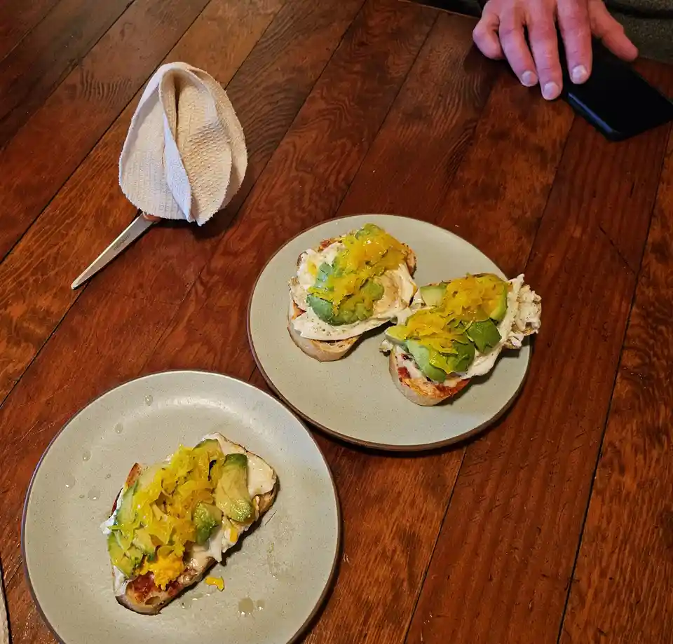 A wooden table holds three plates of avocado toast topped with yellow pickled onions and a fried egg. A hand rests on a…