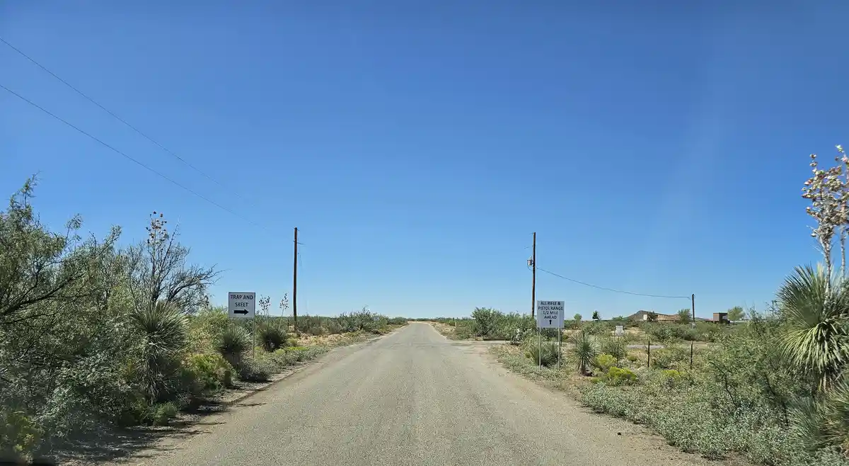 A desert road flanked by cacti and shrubs under a clear blue sky, with two signs: one pointing right for 'TRAP AND SKEET'…
