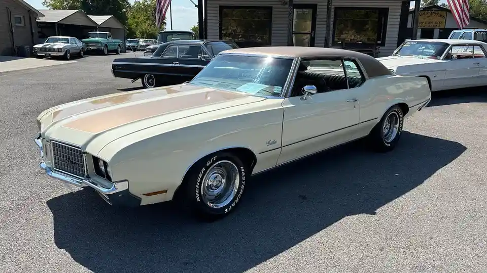 A cream-colored 1970s Oldsmobile Cutlass with a brown vinyl roof and chrome trim is parked in a used car lot under bright…