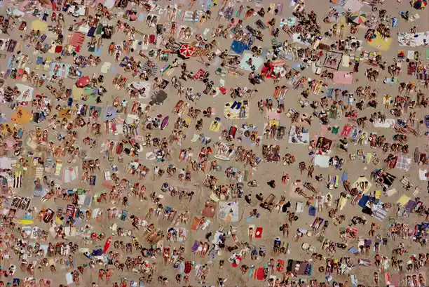 An aerial view of a densely packed beach filled with countless people sunbathing on colorful towels and under umbrellas…