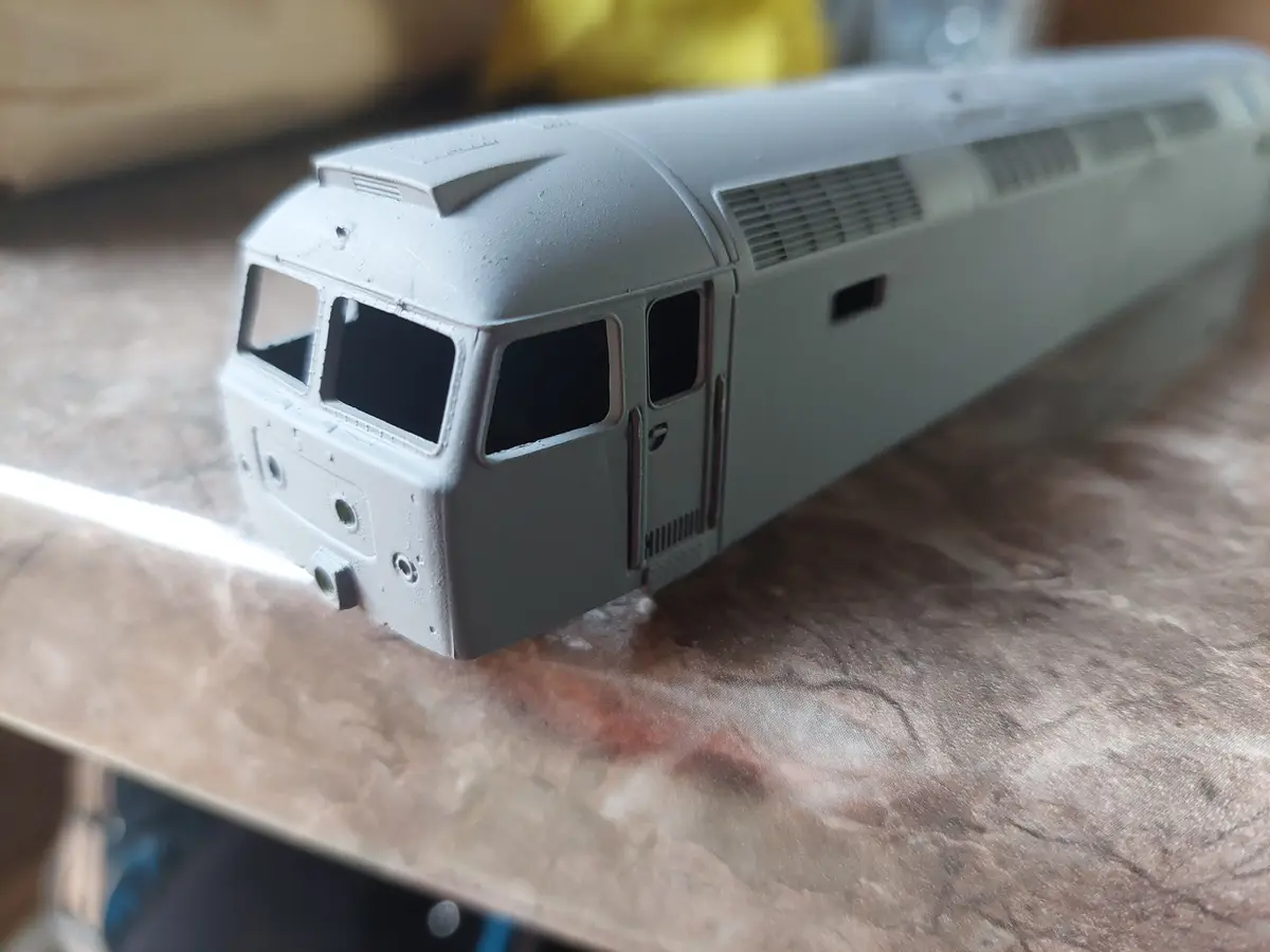 A close-up photo shows a grey-painted model train locomotive on a wooden table, with visible spray paint overspray on the…