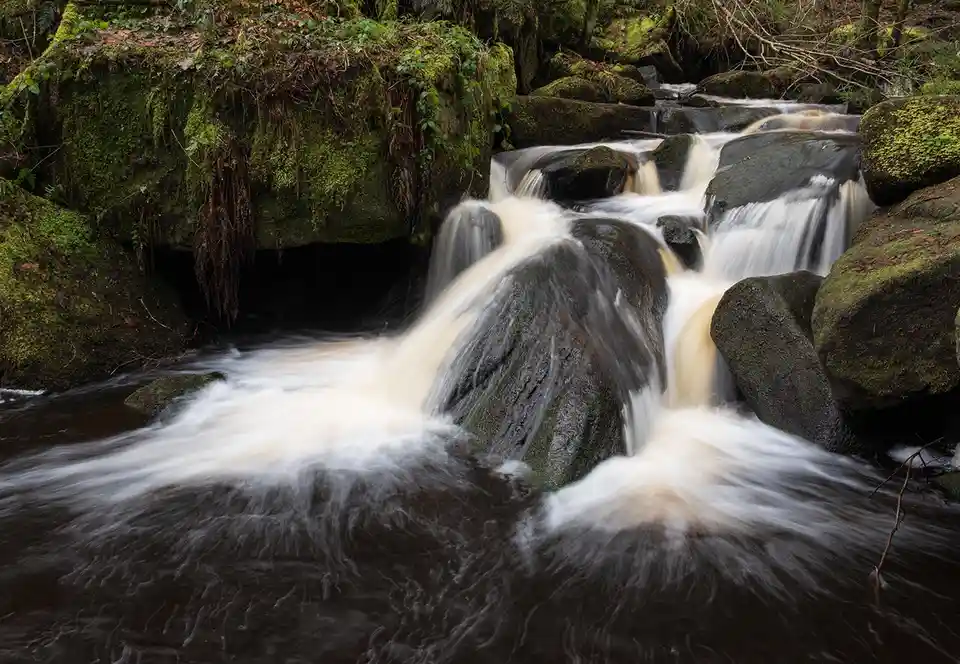 A long-exposure photograph of a moss-covered rocky stream with smooth, flowing water cascading over dark stones. The image…