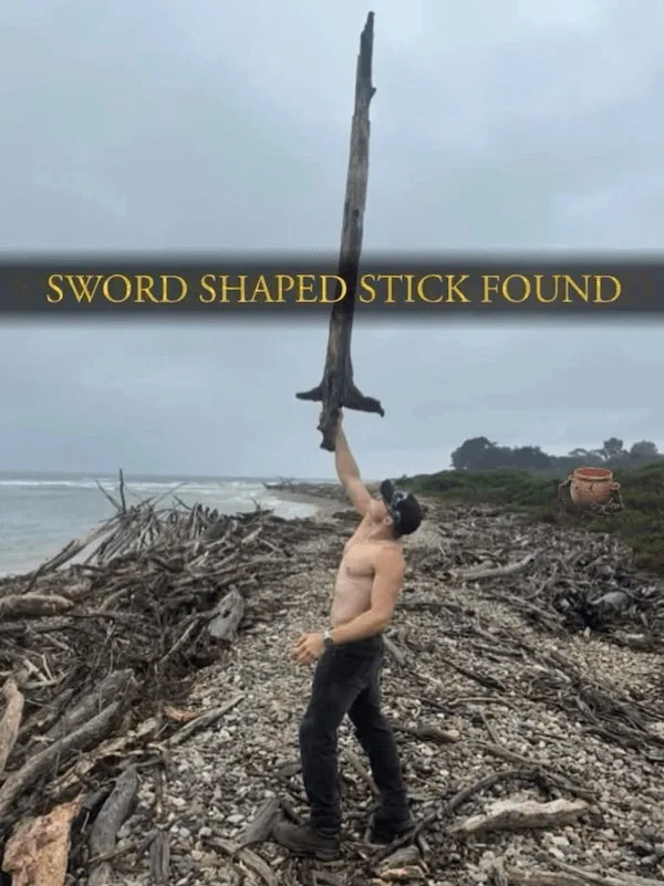 A shirtless man stands on a pebbly beach covered in driftwood, holding up a long, weathered stick that vaguely resembles a…