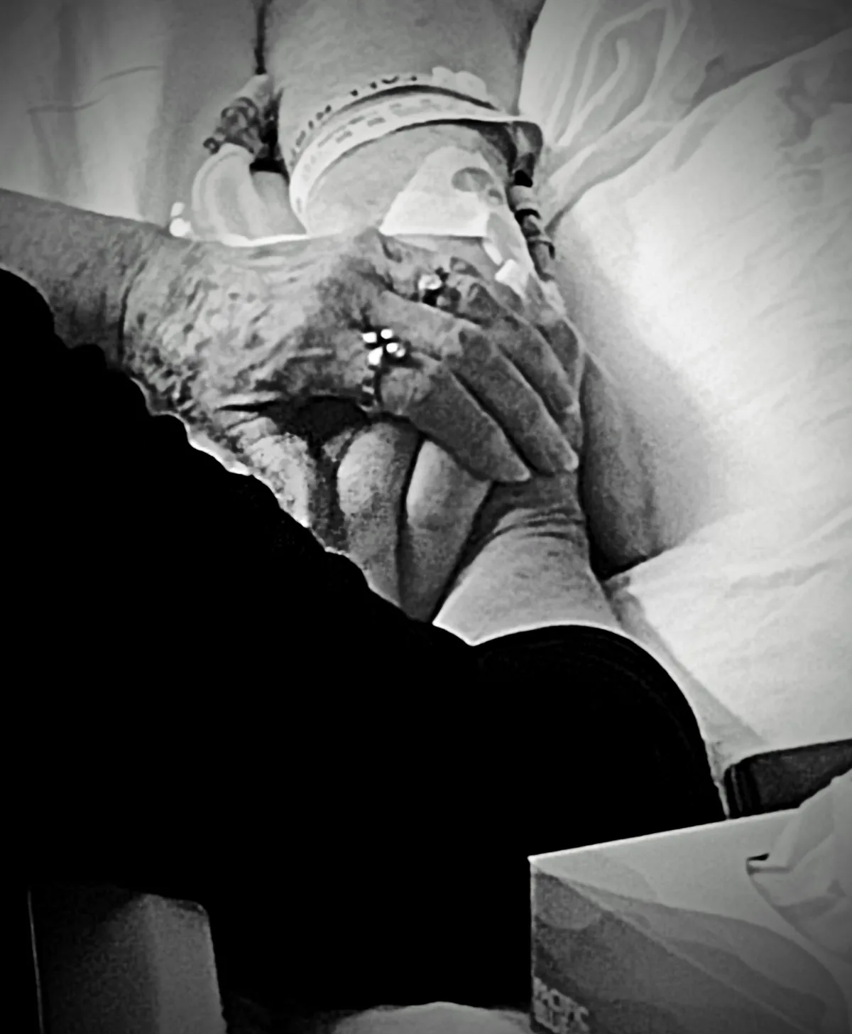 A black-and-white photo shows a person in a hospital bed with an IV and medical tape, holding another person's hand. The…