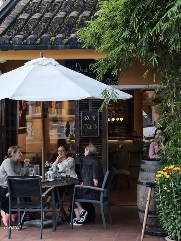 Three women sit at an outdoor café table under a white umbrella, with a small chalkboard sign reading 'we're open' visible…