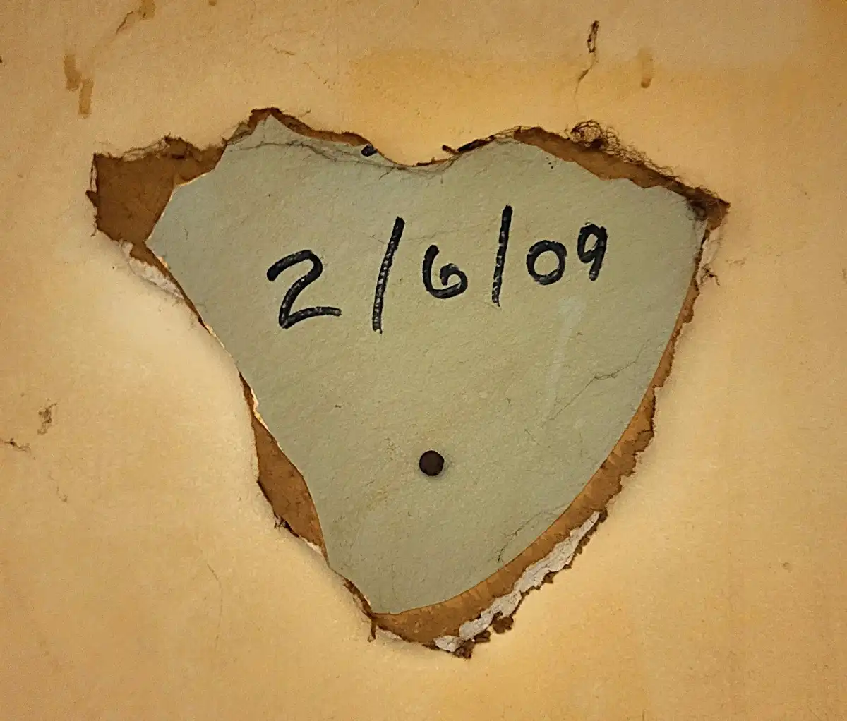 A close-up of a roughly heart-shaped hole punched through a beige wall, revealing a lighter green inner layer. The date…
