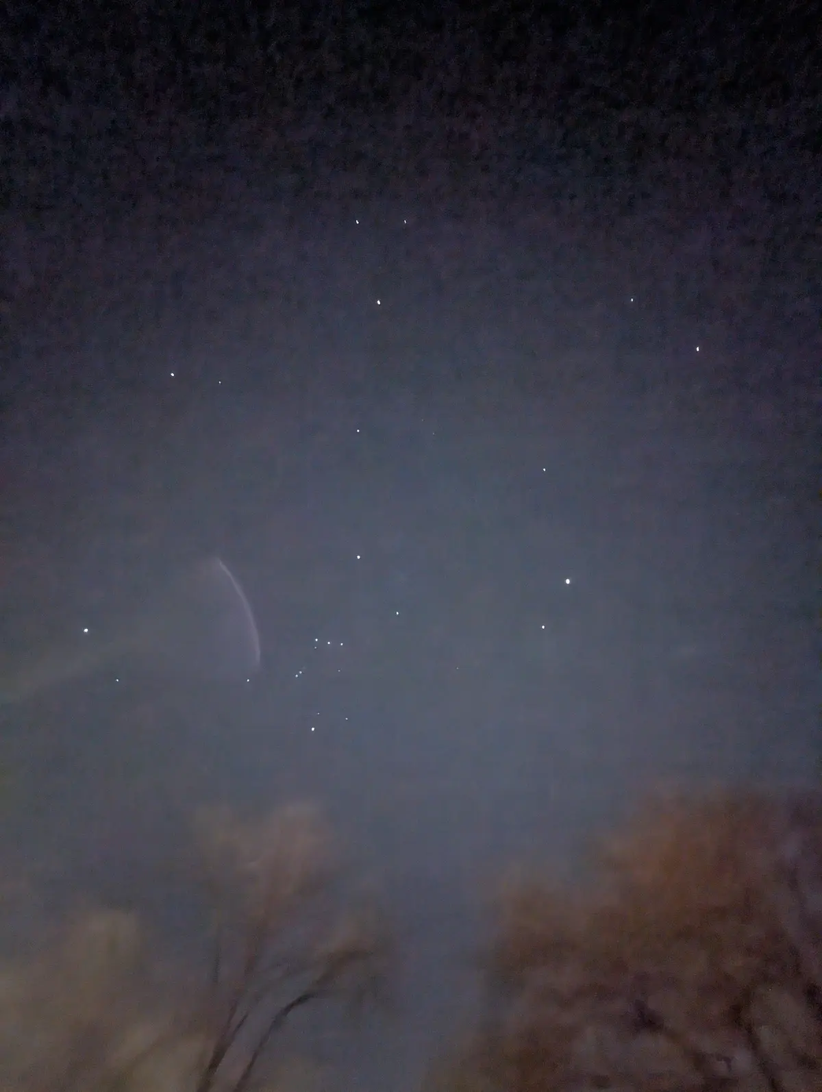 A blurry night sky photo showing a faint crescent moon partially obscured by haze or clouds, with scattered stars visible…