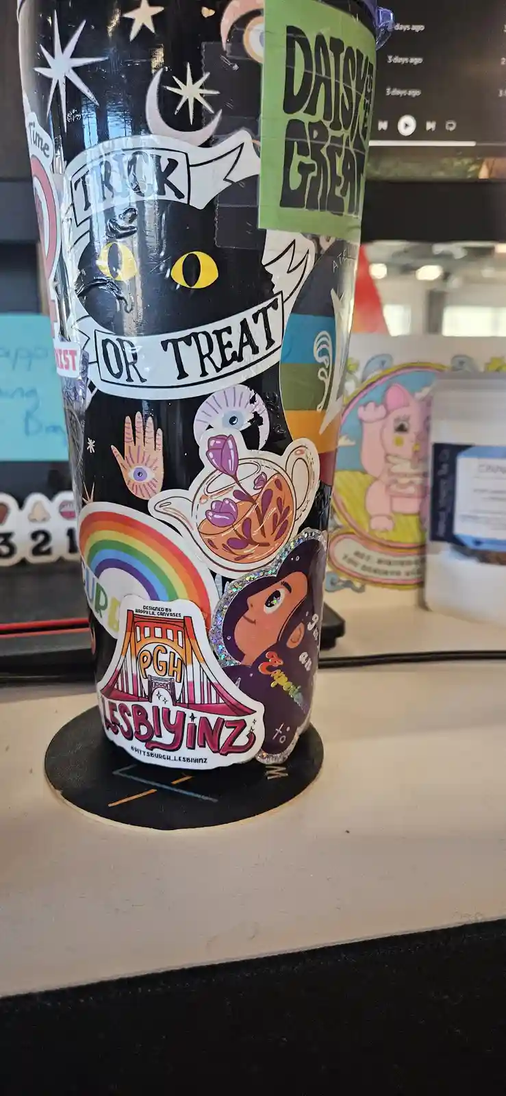 A black tumbler covered in colorful stickers, including a large black cat with yellow eyes under a 'trick or treat' banner…