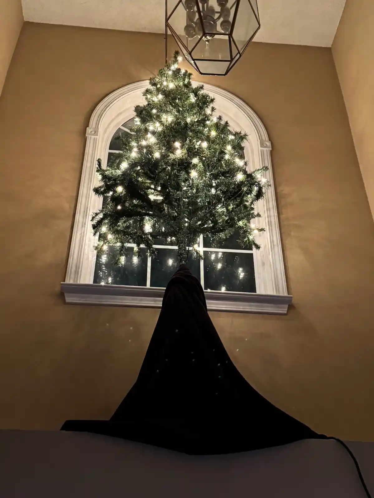 A decorated christmas tree with lights is positioned inside an arched window, its lower half covered by a dark cloth draped…