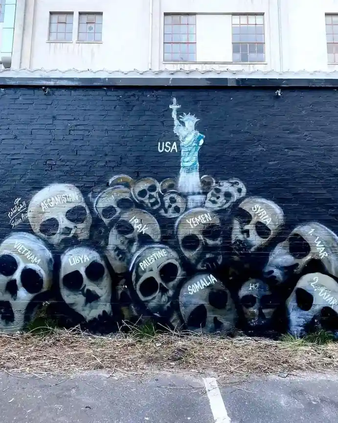 A black brick wall mural depicts a blue Statue of Liberty labeled 'USA' standing atop a pile of skulls, each labeled with a…