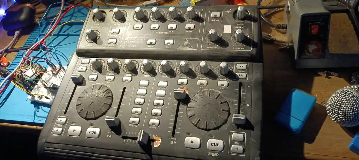 A close-up photo of two black Behringer DJ controllers on a cluttered wooden desk, surrounded by wires, a soldering iron…