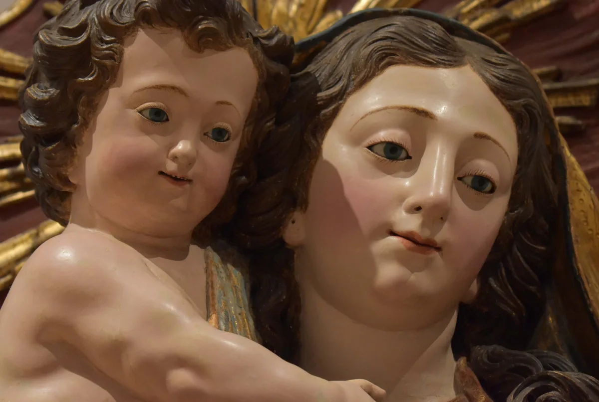A close-up of a classical religious statue showing the Virgin Mary holding the infant Jesus. The baby Jesus looks directly…