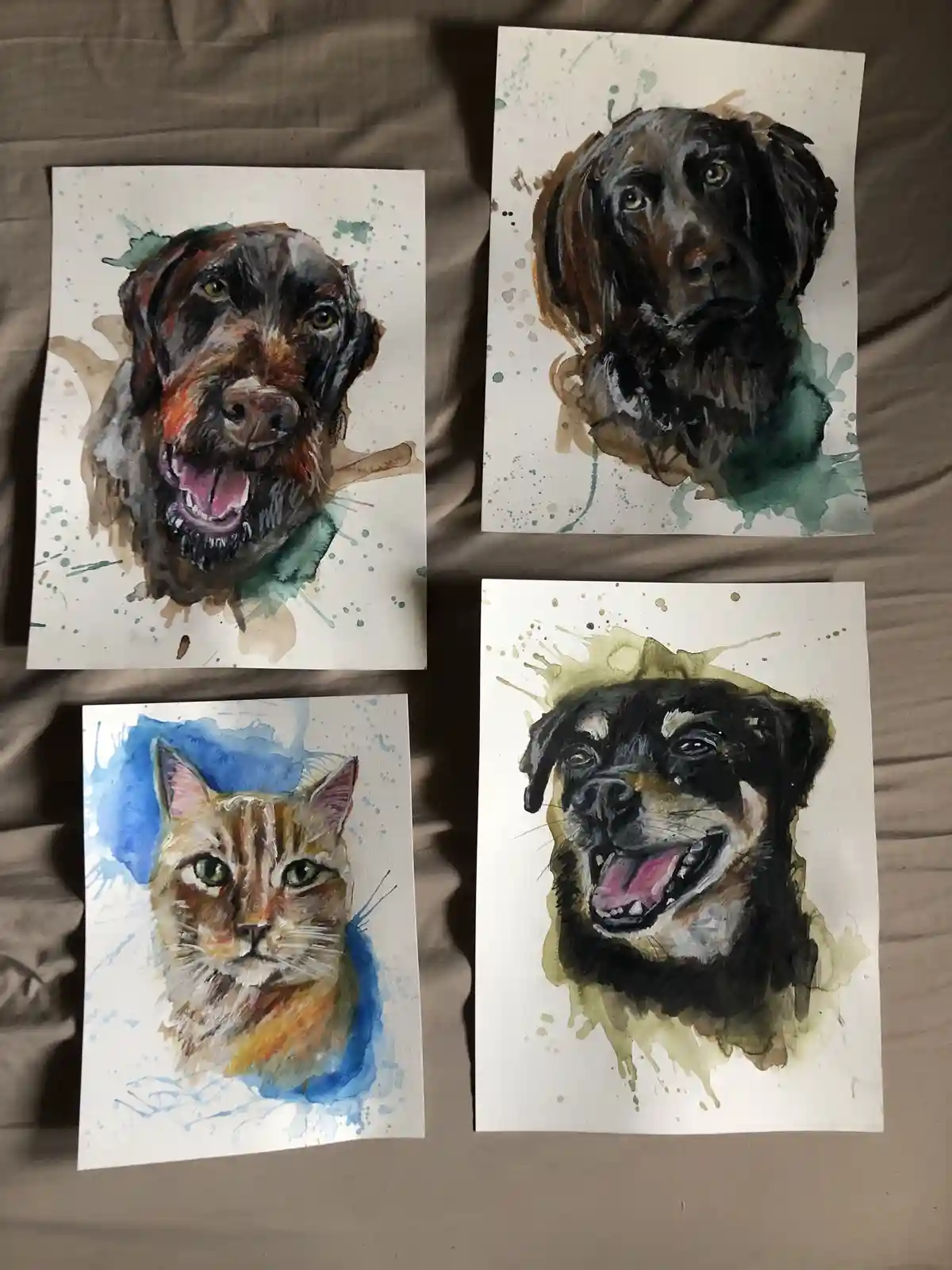 Four watercolor pet portraits are laid out on a brown fabric surface. Three depict dogs with expressive faces—one with an…
