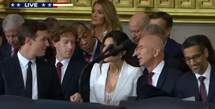 A group of people, including Jeff Bezos, are seated at a press conference. Bezos is speaking into a microphone while others…
