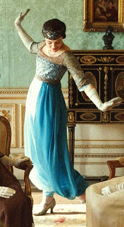 A woman in a vintage blue and white dress stands in an ornate room, arms outstretched as if dancing, but her feet are…