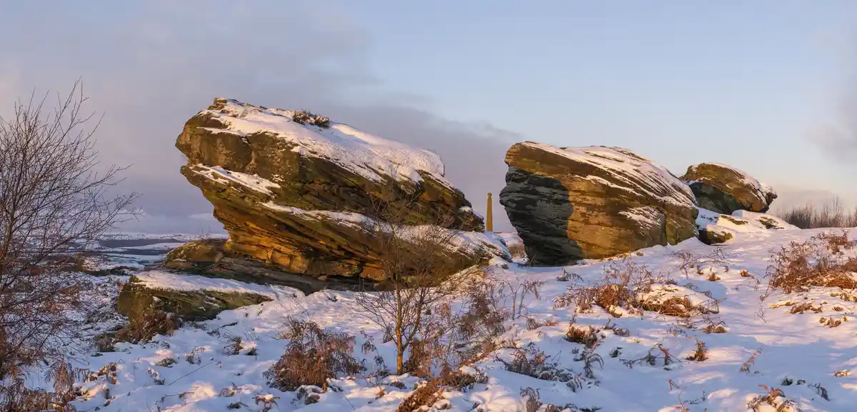 A snowy landscape with large, layered rock formations under a pale sky, with bare bushes and a thin layer of snow covering…