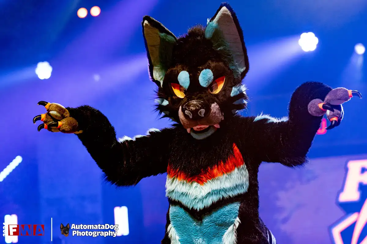 A person in a detailed black, blue, and red fursuit resembling a wolf or dog stands on a stage under blue lights with arms…