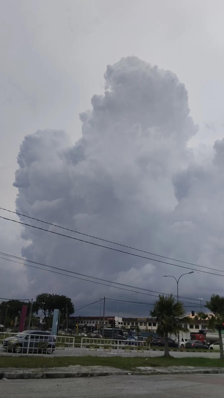 A large, ominous, vertically stacked cloud formation dominates the sky over a street scene with cars, palm trees, and…