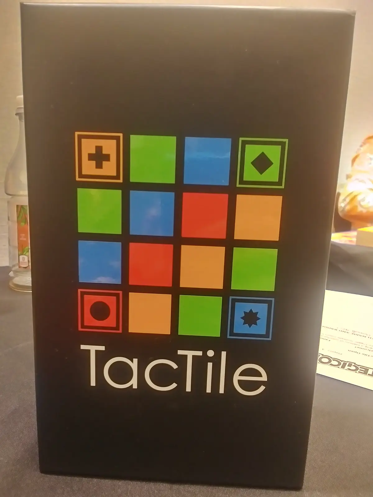 A black game box for 'TacTile' features a 4x4 grid of colorful squares with various symbols like a plus sign, diamond…