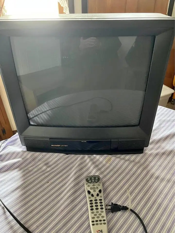 A black Sharp Inten CRT television sits on a striped bedsheet, its screen off and reflecting a person taking the photo. In…