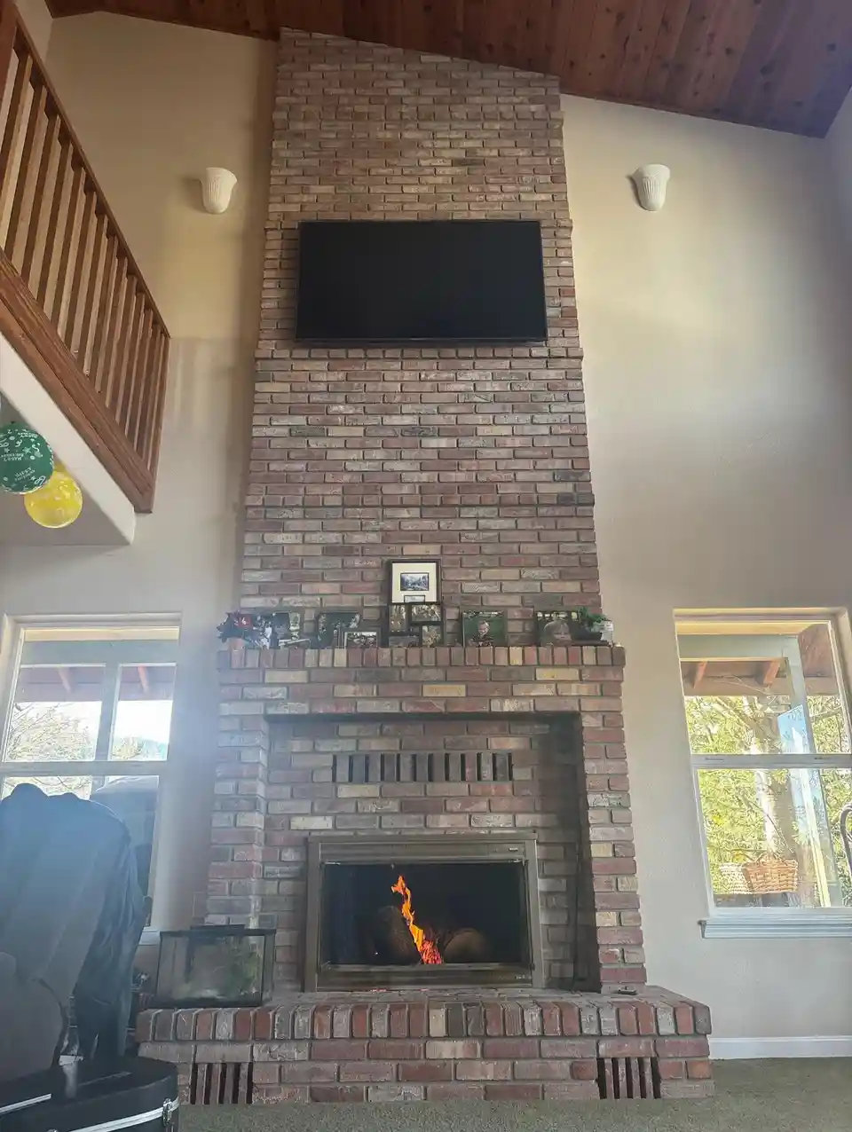 A photo of a tall brick fireplace with a fire burning inside, a TV mounted above it, and family photos on the mantel. The…