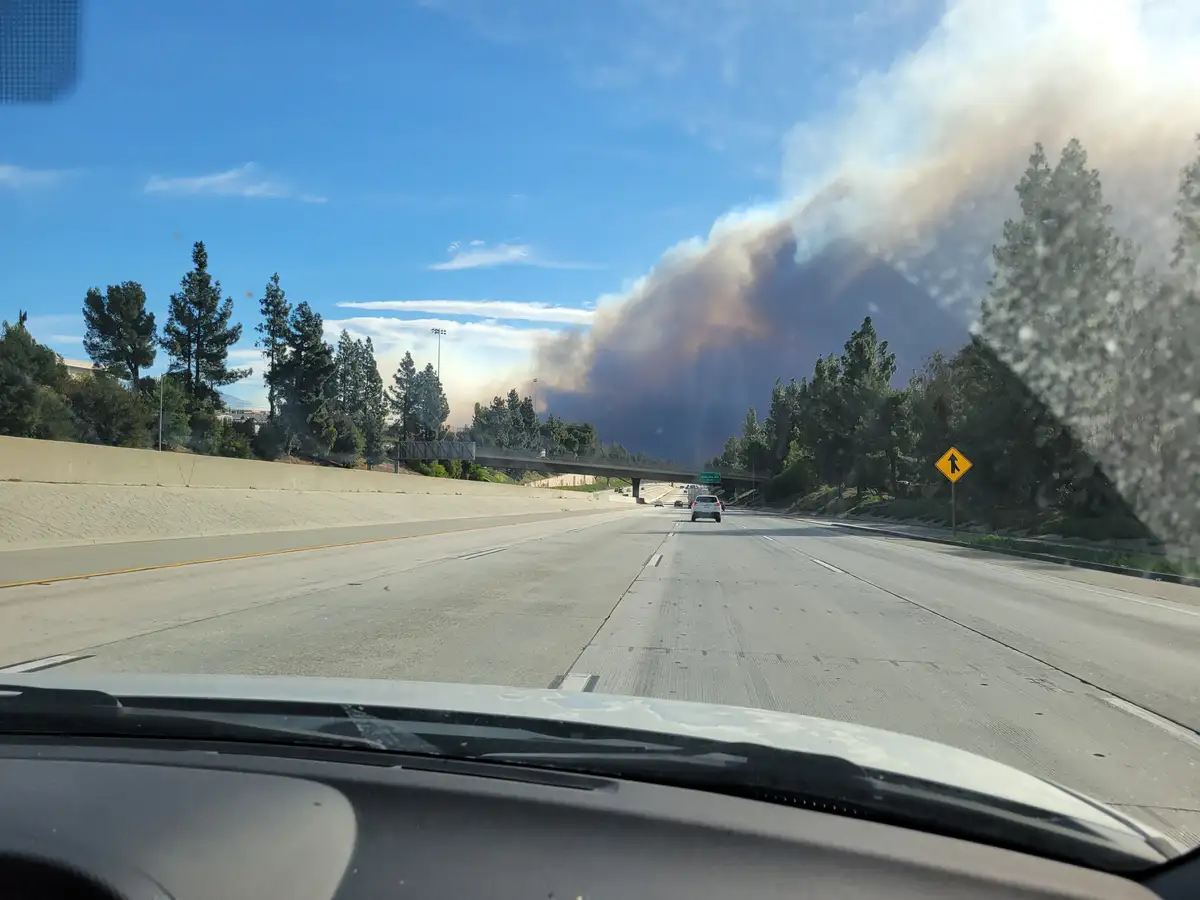 A photo taken from inside a car on a highway shows a massive plume of smoke rising from a forested hillside under a blue…
