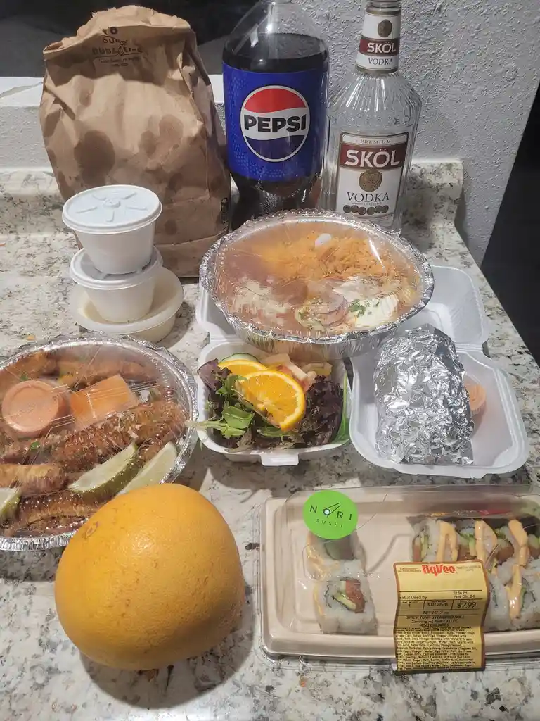 A countertop displays a large, messy spread of takeout food including sushi, fried items, salad, and a whole grapefruit…