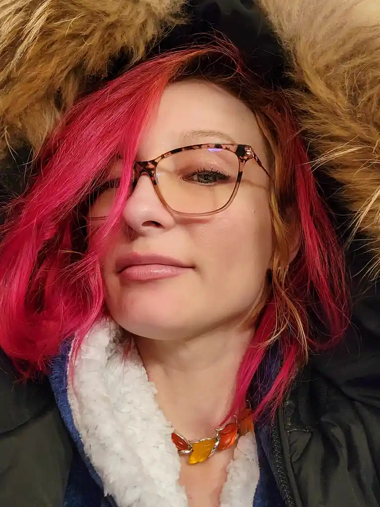A person with bright pink hair and tortoiseshell glasses wears a dark winter coat with a fluffy brown fur-lined hood. They…