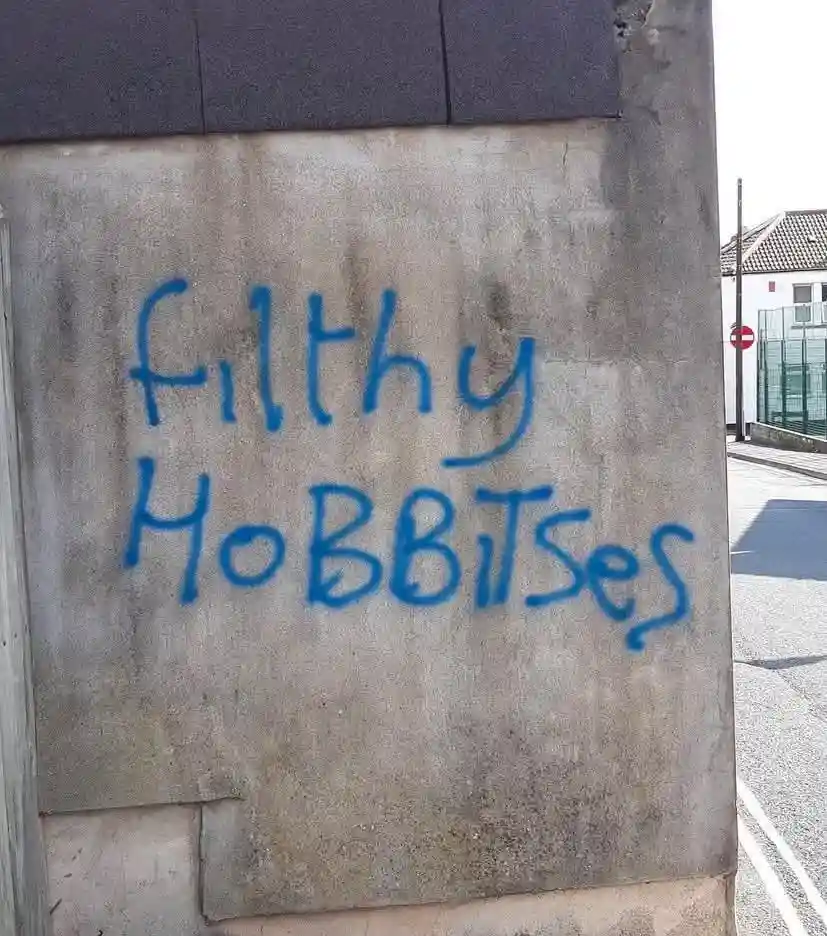 Blue spray-painted graffiti on a concrete wall reads 'filthy HOBBITsEs' in a sloppy, all-caps style. The wall is part of an…