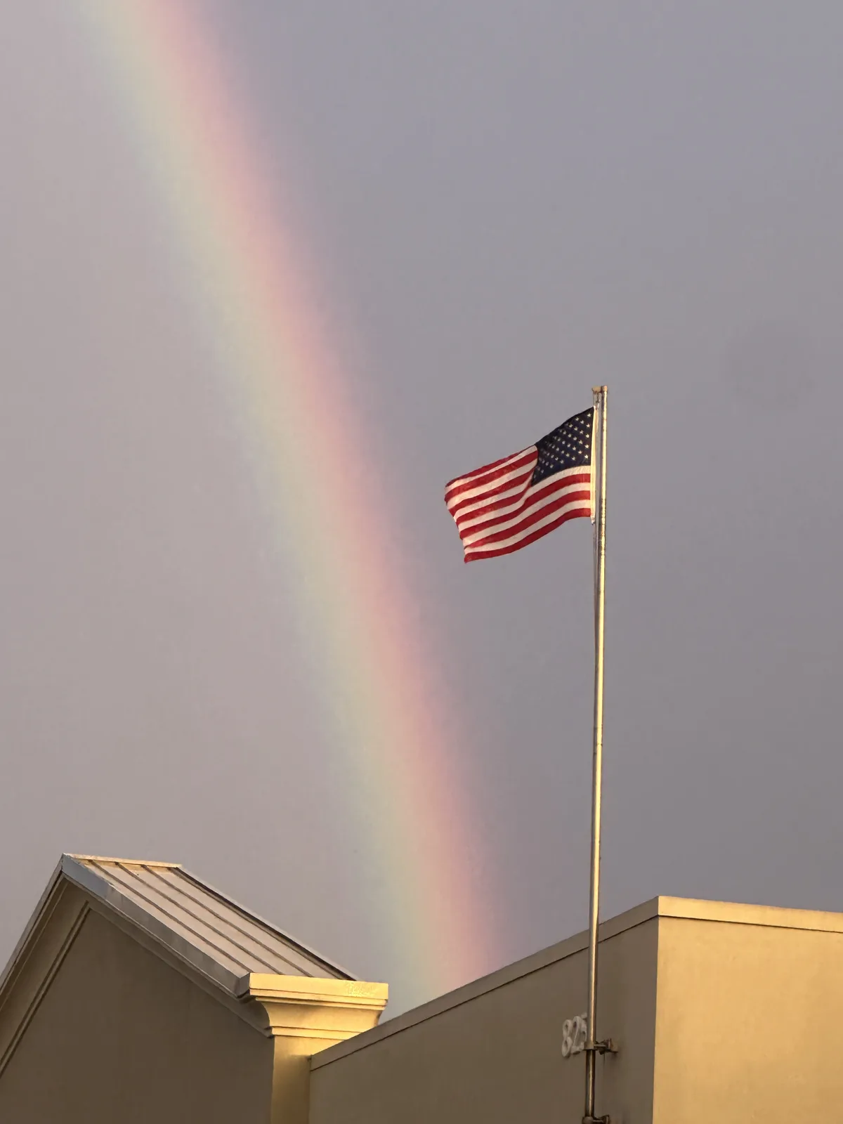 An american flag flies atop a building against a gray sky, with a faint rainbow arcing behind it. The overlaid text reads…