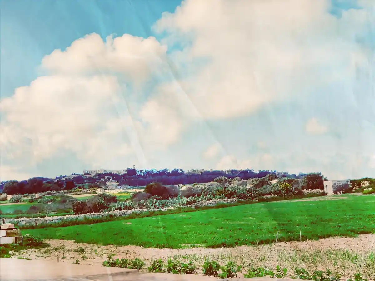 A vintage-style landscape photo shows a green field with stone walls, distant buildings, and a cloudy sky with visible light…