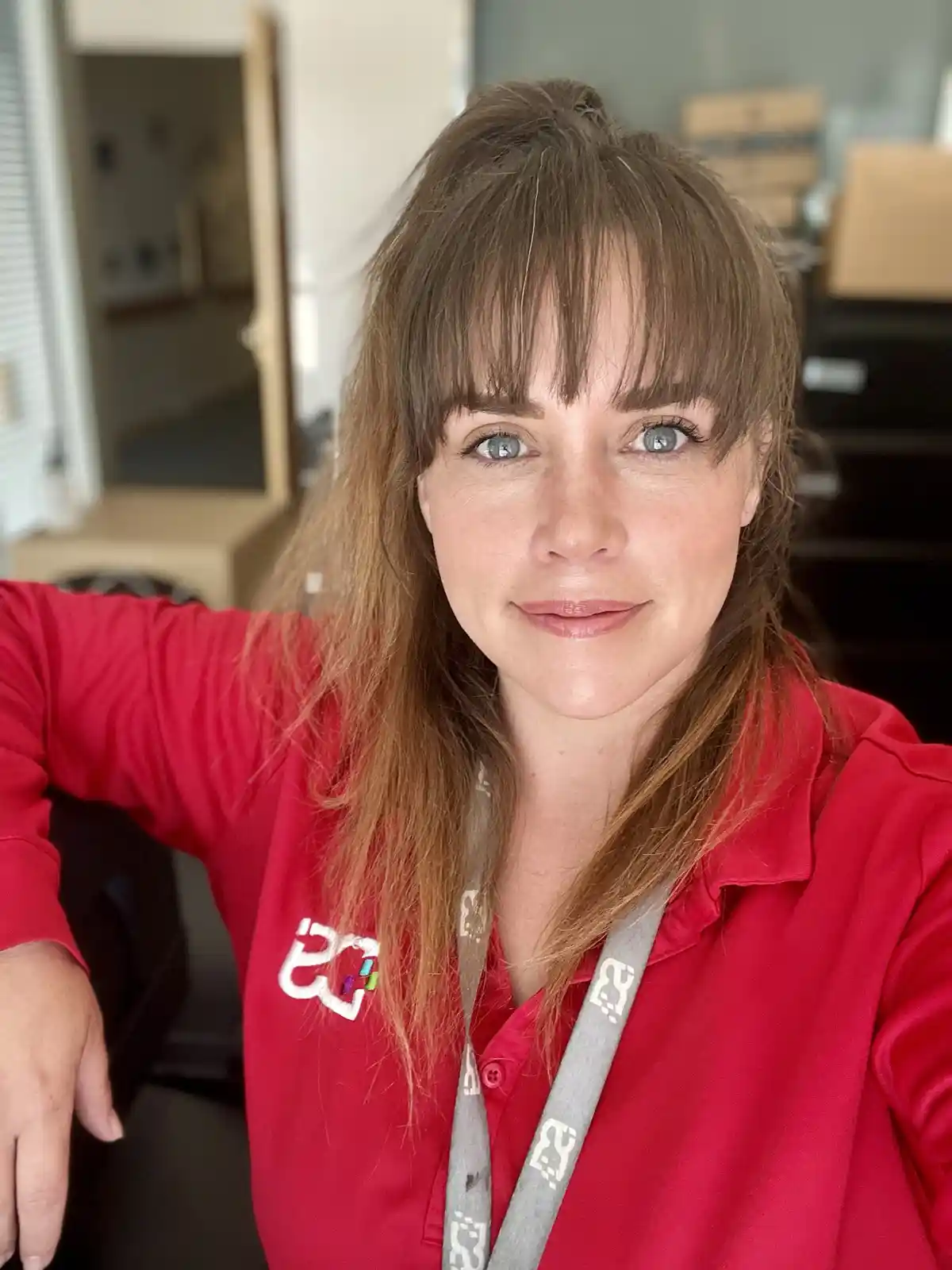 A woman with brown hair and blue eyes wearing a red polo shirt and a grey lanyard takes a selfie. The overlaid text reads…