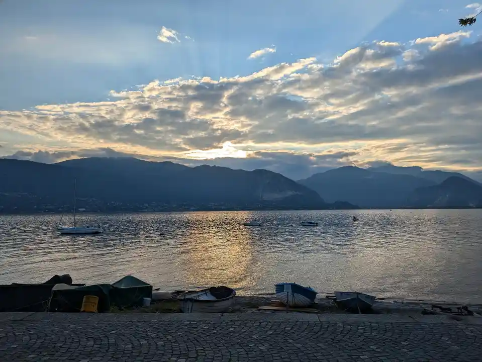 A serene lakeside scene at sunset with mountains in the background, boats on the water and shore, and a cobblestone path in…