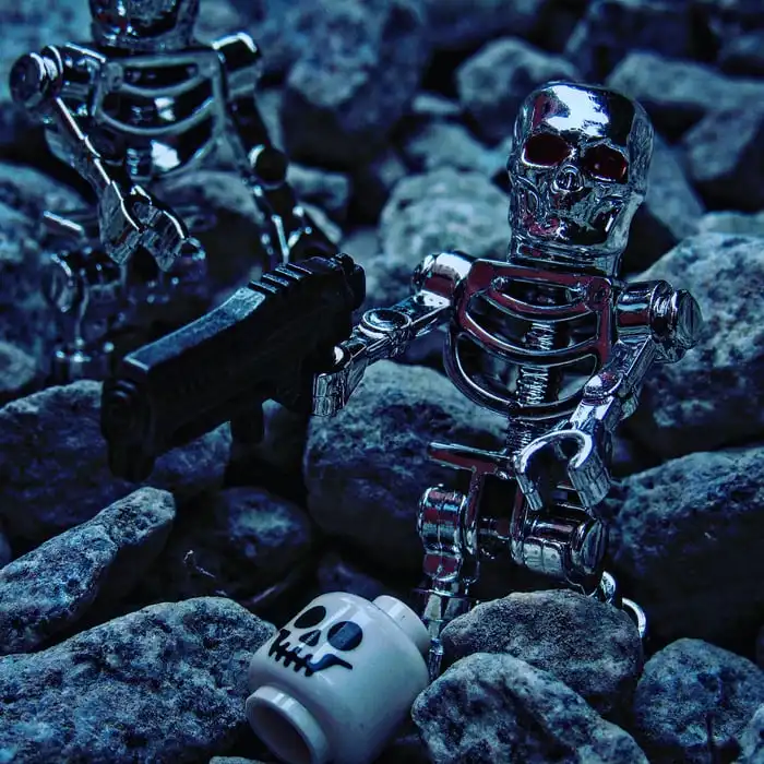 Two chrome skeleton robot figures, one holding a gun, stand among dark rocks. A white lego head with a skull face lies…