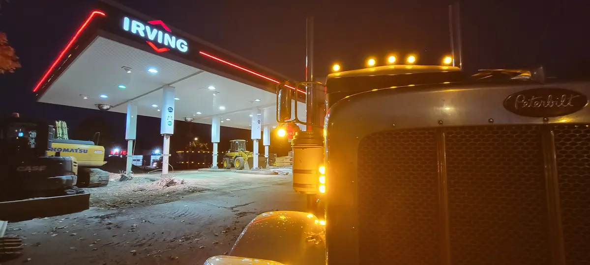 A nighttime photo shows the front of a Peterbilt semi-truck with its amber lights on, parked at an Irving gas station. The…