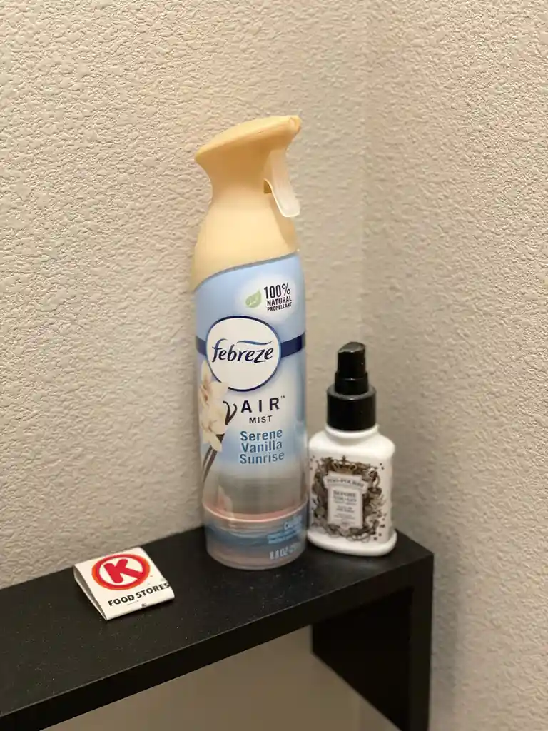 A photo shows a black shelf against a textured wall holding a large Febreze Air Mist spray bottle labeled 'Serene Vanilla…