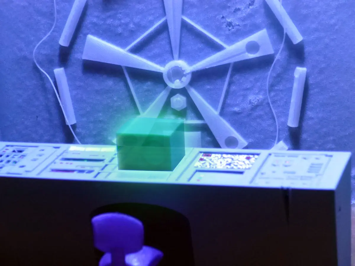 A close-up of a 3D printer's build platform under blue light, showing a partially printed, translucent green cube in the…
